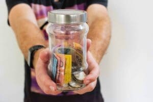 extended arms with hands clasping a glass jar, with a silver lid. The Jar contains various Australian currency notes and coins.