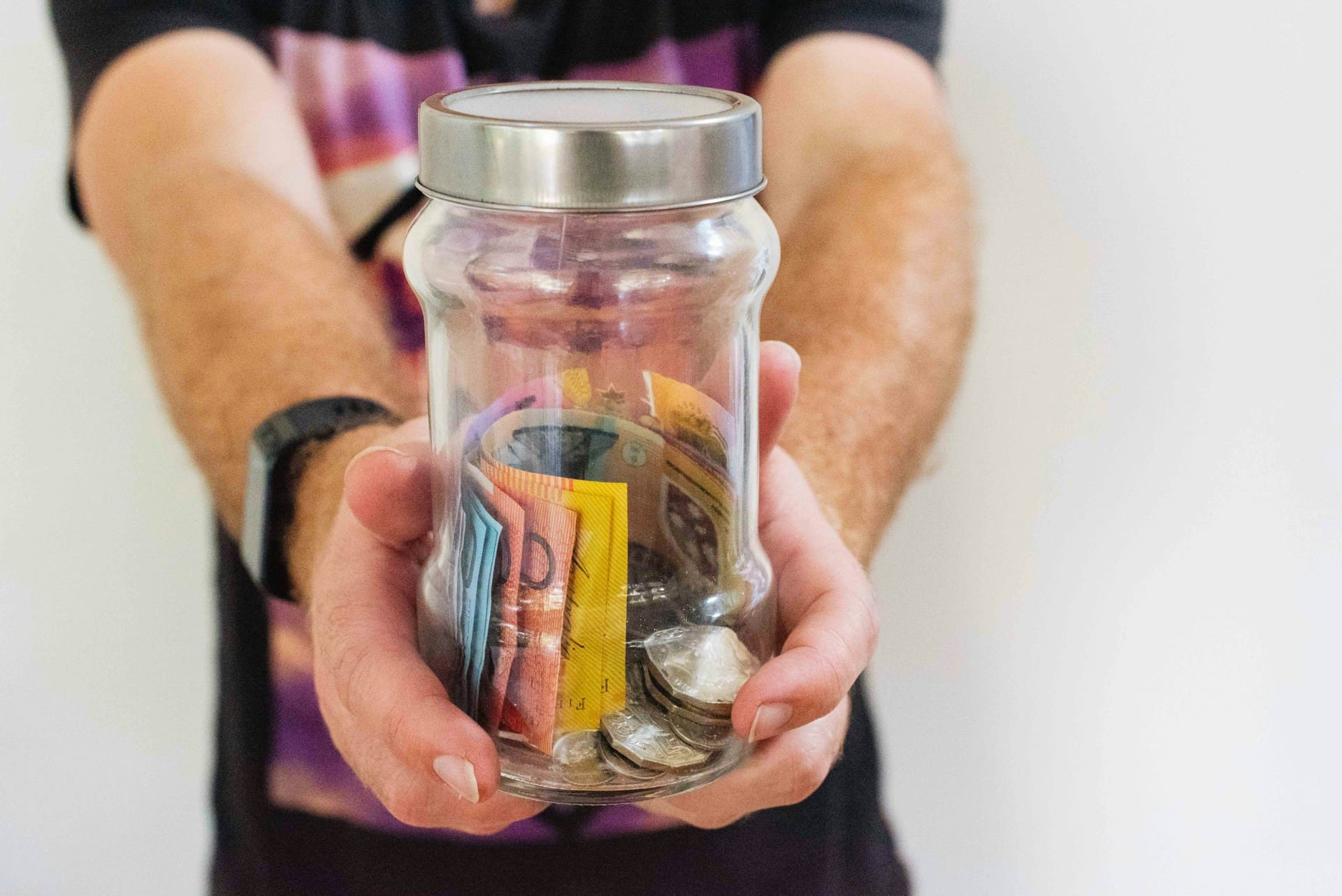 extended arms with hands clasping a glass jar, with a silver lid. The Jar contains various Australian currency notes and coins.